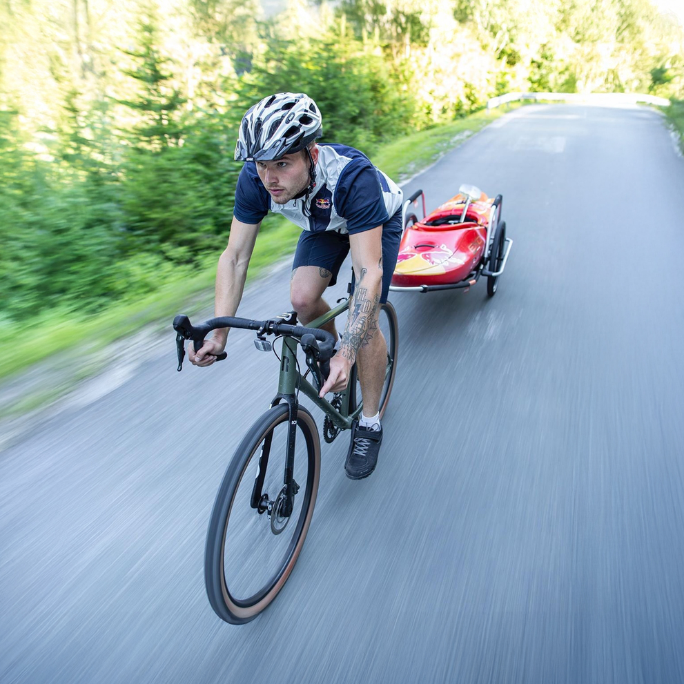 A cyclist riding on a forest road with a Reacha Sport bike trailer carrying a red kayak, highlighting its stability on outdoor terrain.