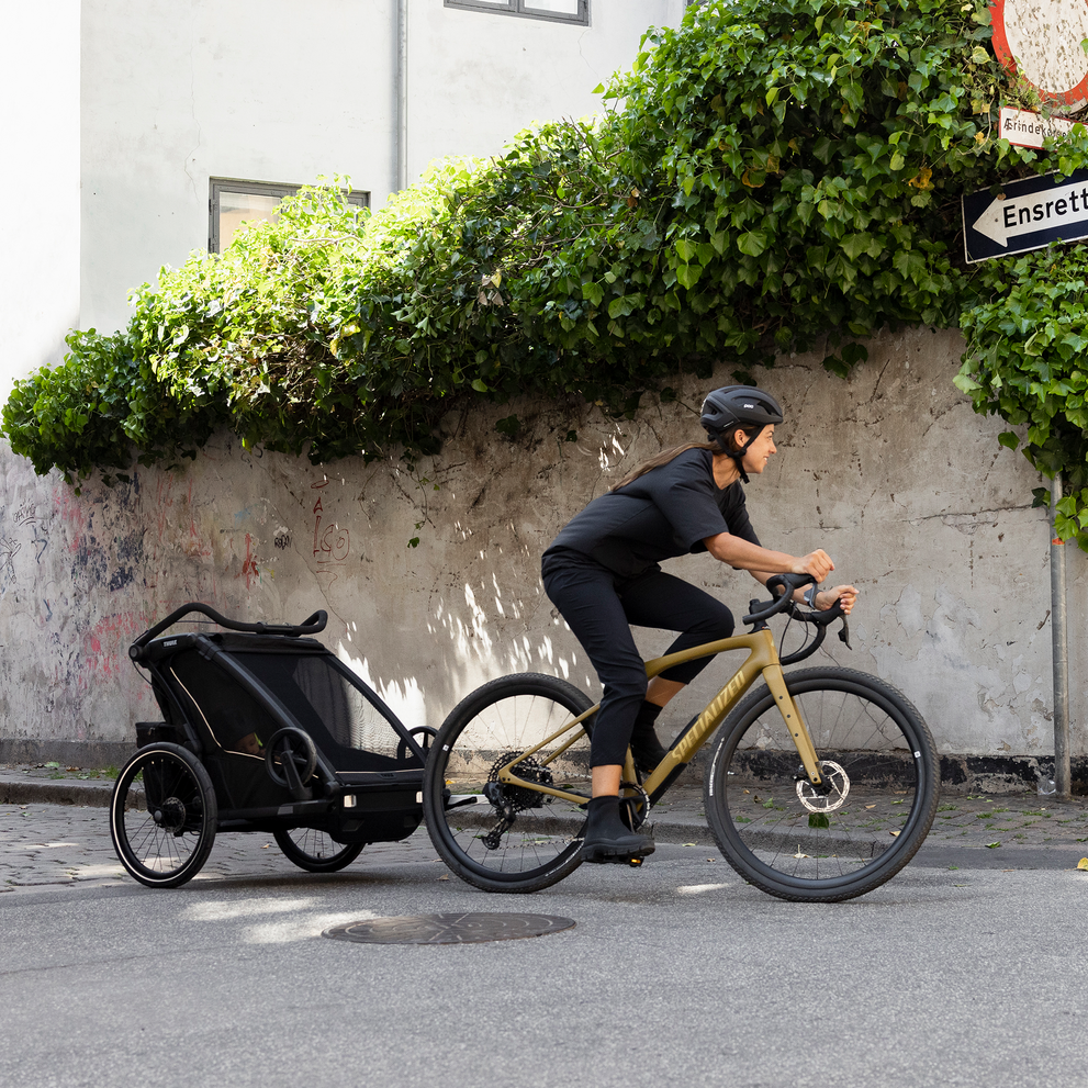 A woman bikes down a sunny street with her black Thule Chariot Sport kids bike trailer.