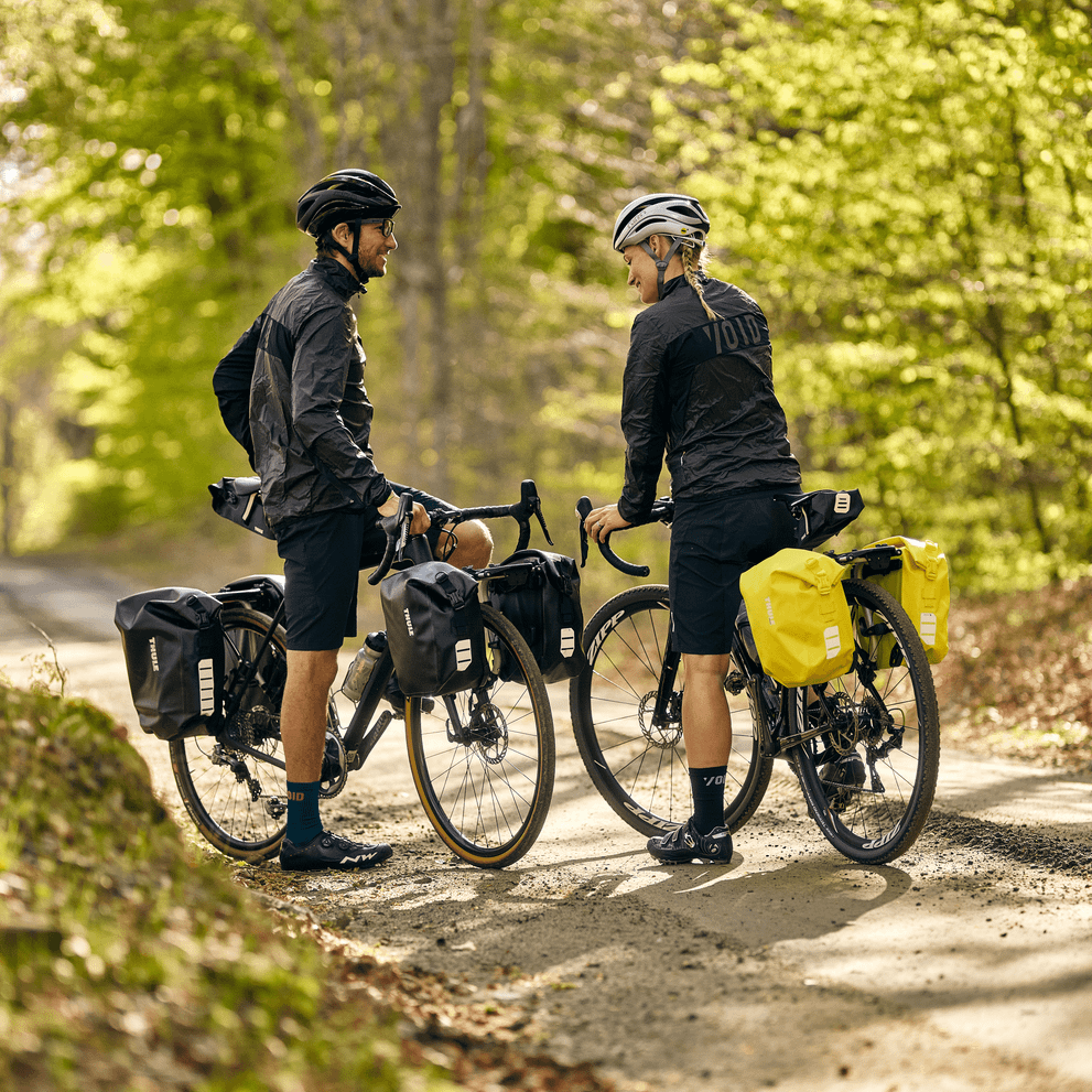 Two cyclists stand on a trail wearing black, carrying Thule Shield Pannier bike bags.