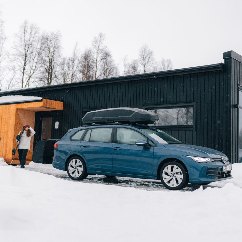 A blue car parked outside a modern black wooden house in a snowy landscape. The car is equipped with a Thule Force 3 roof box and a Thule Onto 2 cargo carrier mounted on the towbar. A person is seen near the entrance of the house, dressed for winter.