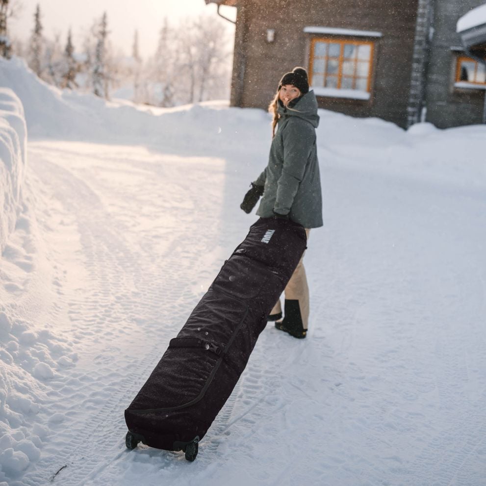 A woman stands in the snow outside a cottage wheeling her Thule RoundTrip travel ski bag.