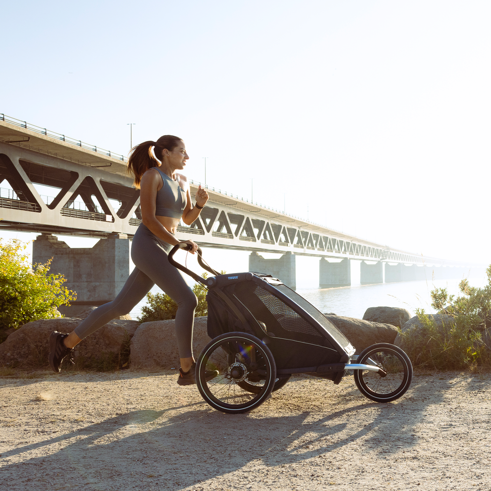 A woman runs by a bridge with her Thule Chariot Sport jogging bike trailer.