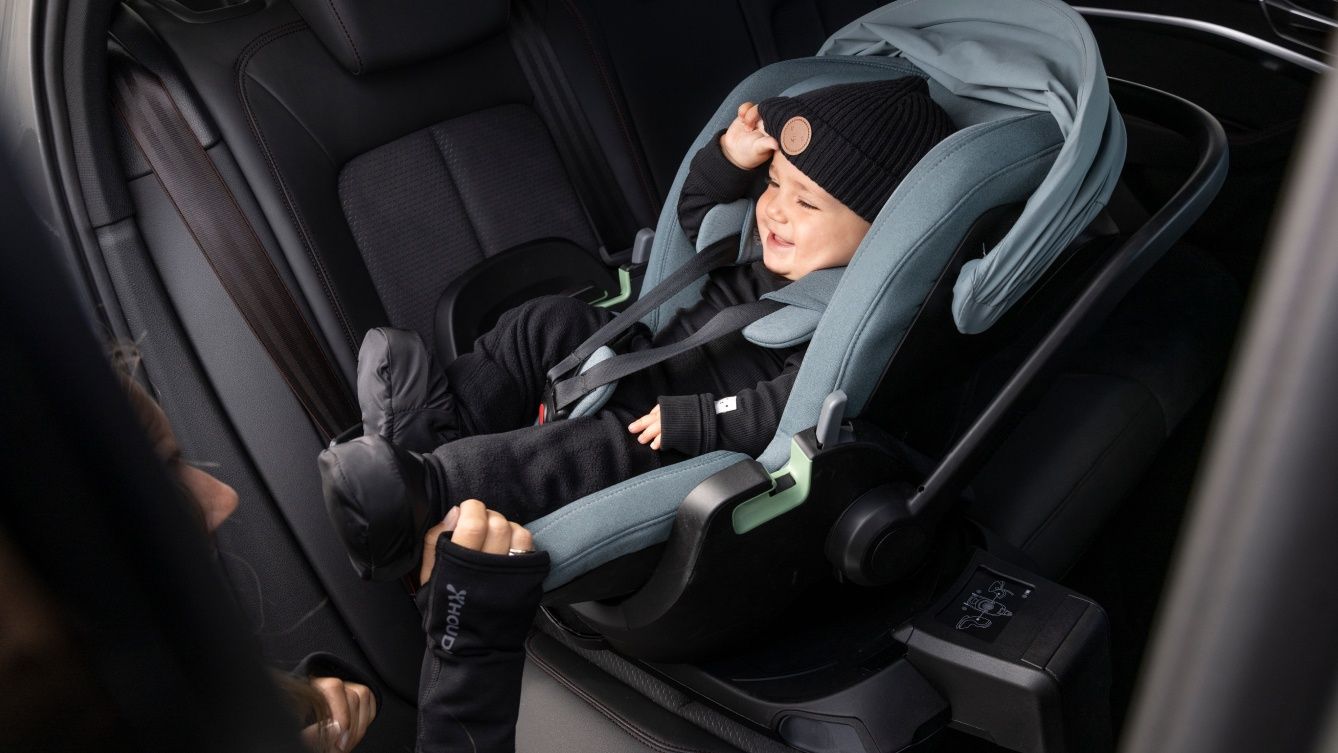 A baby smiling in a Thule Maple infant car seat mounted on the base. The seat is turned outwards and a person’s hands are touching the edge of the car seat.