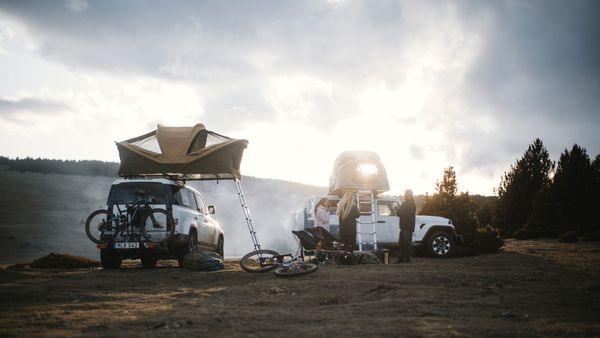 Un acogedor campamento al anochecer en un paisaje escarpado, con varios vehículos equipados con tiendas de techo Thule Approach y un grupo de personas.