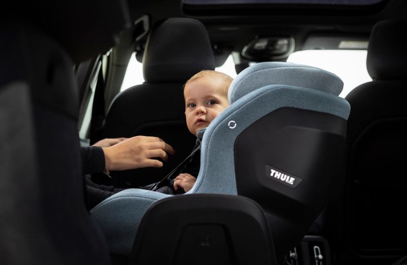 A young toddler sitting in a Thule Elm toddler car seat in light grey. A person's hands are near the toddler's straps.