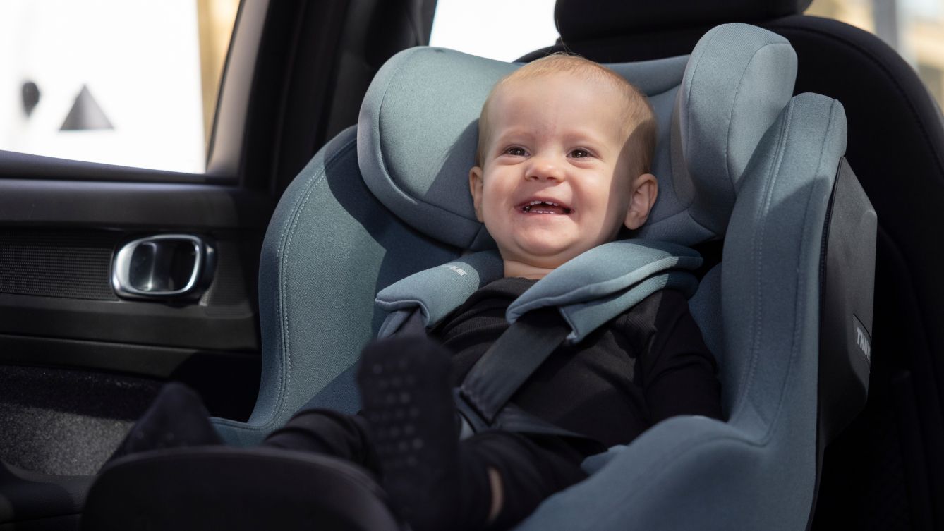 A baby smiling wide while sitting in a mid-blue Thule Maple infant car seat, mounted on the back seat.