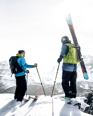 Zwei Personen stehen mit Ski und Skirucksäcken auf einem Berg.
