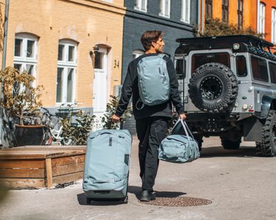 A man walking down the street with a light blue Thule Chasm wheeled duffel, carrying a matching duffel bag and wearing a coordinated backpack.