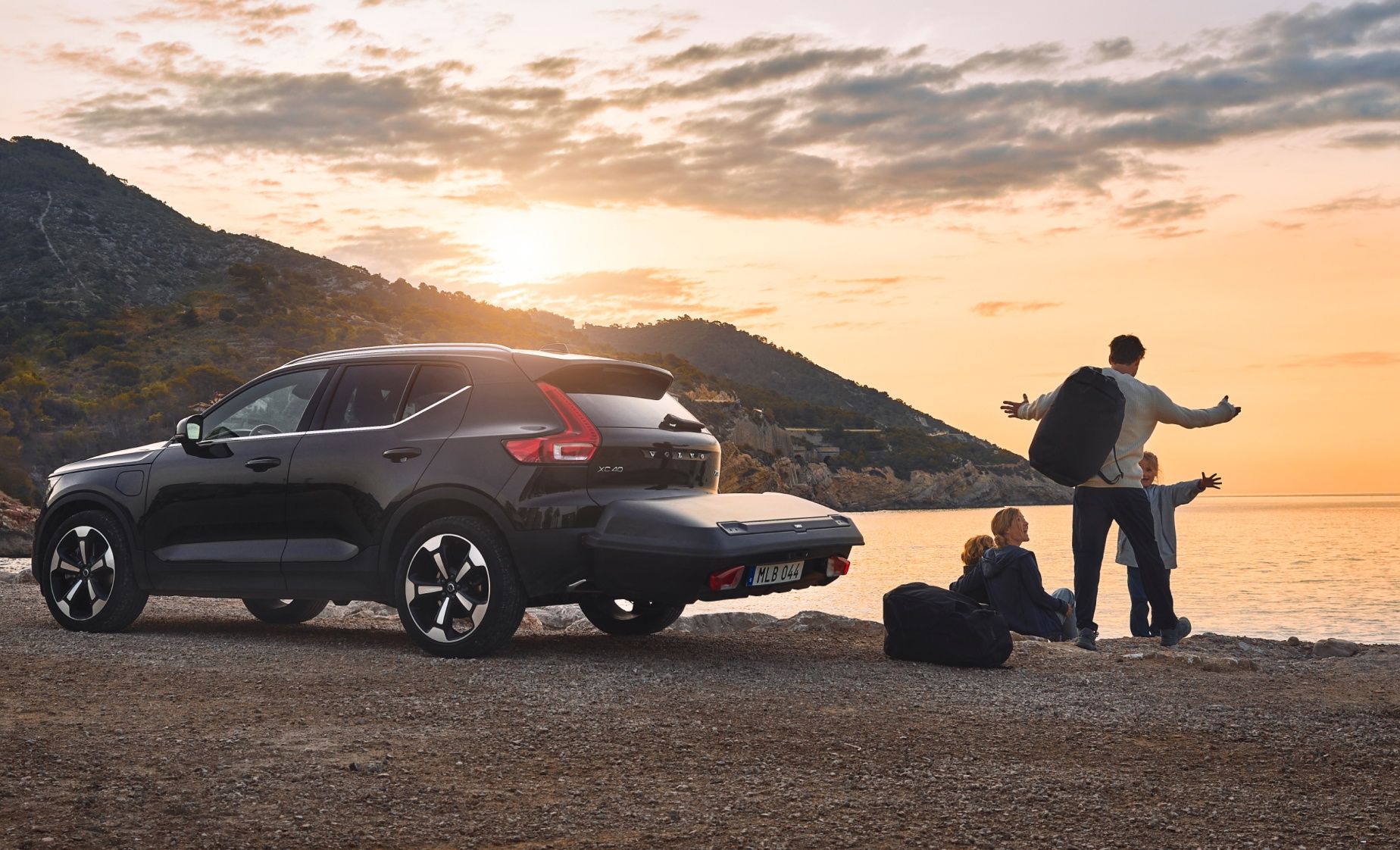 A car is parked next to a beach where a family watches the sun set and a rear cargo box is on the car.