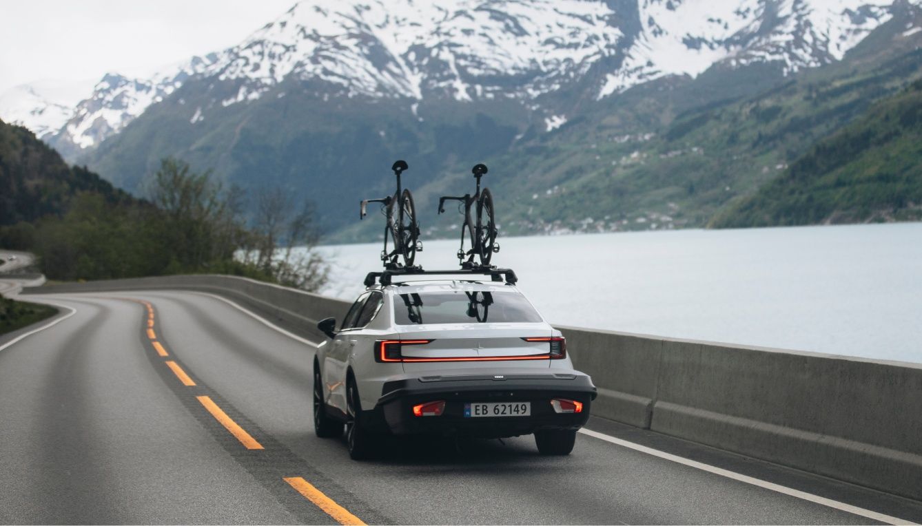 A car drives down a street next to a lake and a mountain with a tow bar box and bikes on the roof.