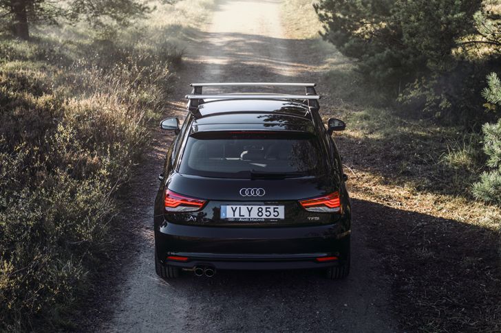 A black vehicle drives down a forest path with roof  racks on the roof.