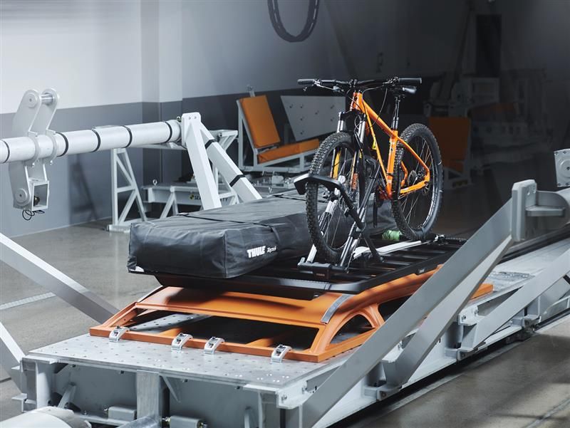 Une installation d’essai de collision comportant un traîneau d’essai doté d’un système de transport de marchandises et de vélos monté à l’arrière Thule. Un vélo de montagne orange vif est solidement fixé au support, ainsi qu’un sac de marchandises noir de marque Thule.