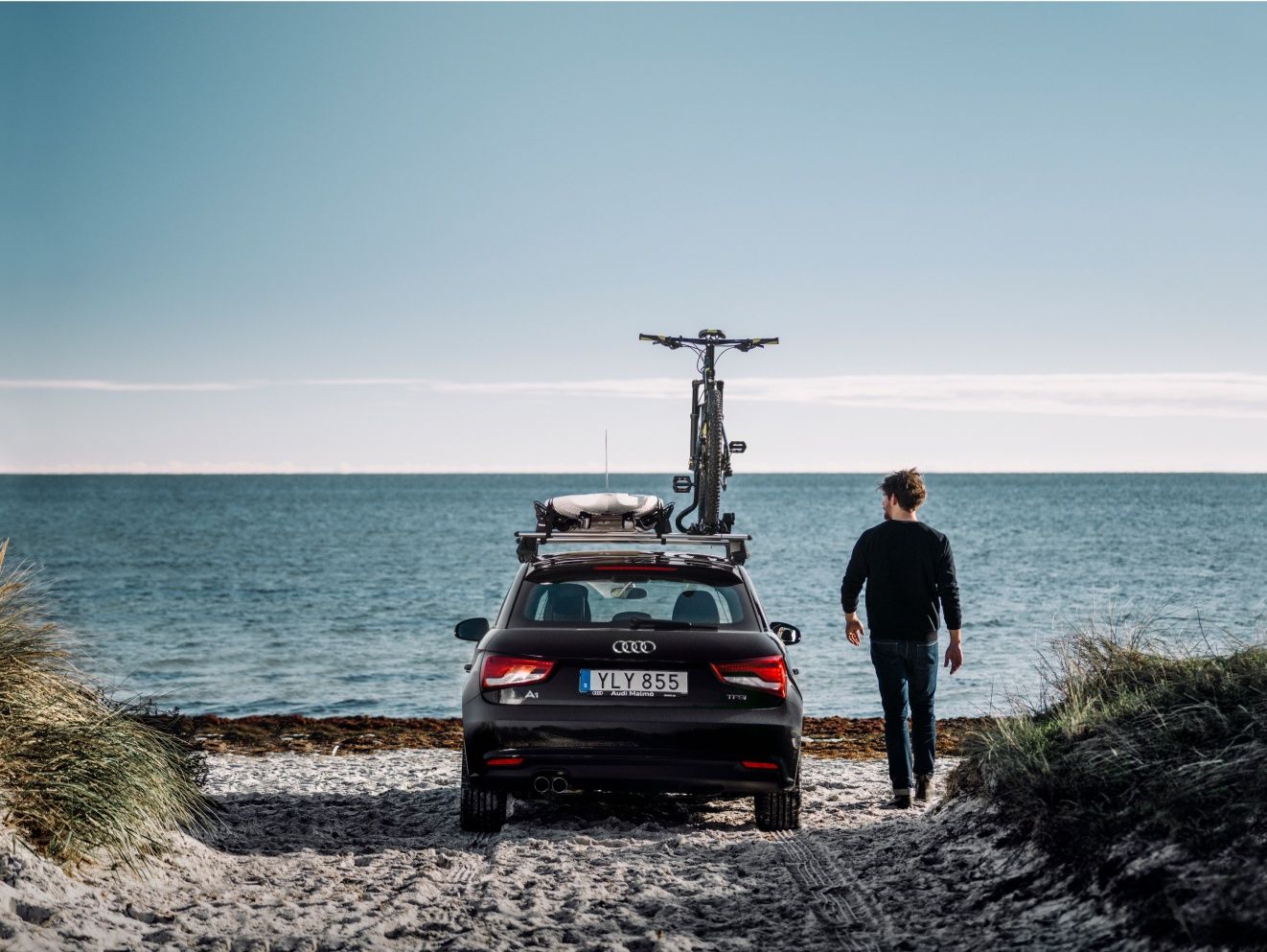 Person standing next to a car equipped with Thule roof racks and bike carrier, parked near the coast, ready for outdoor adventure.