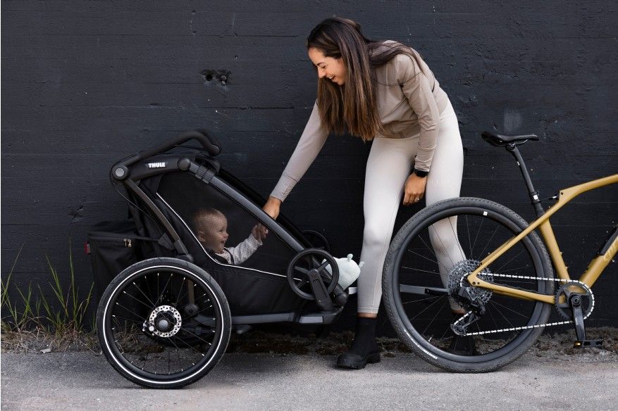 A woman shakes the hand of her daughter in a black bike trailer against a black wall.