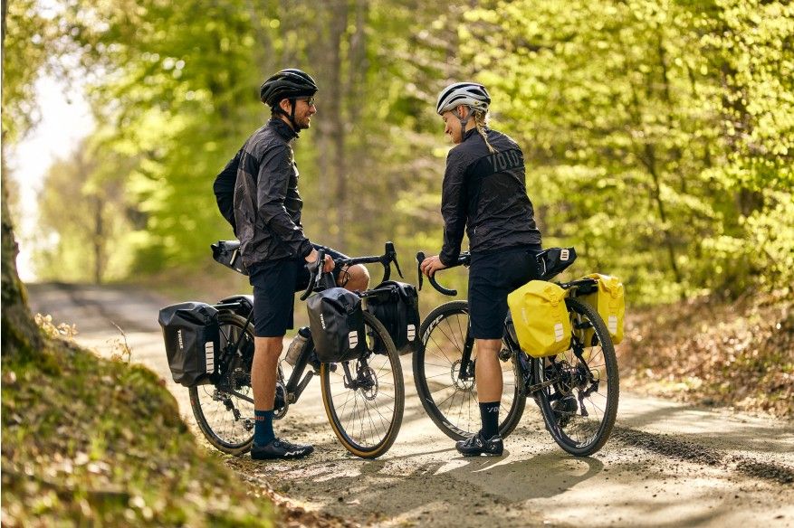 Two cyclists are on their bikes in a forest path with bike packs.