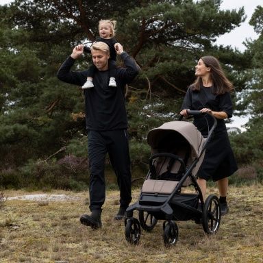Ein Mann geht mit einem Kleinkind auf den Schultern in der Natur neben einer Frau, die einen leeren Thule Urban Glide Kinderwagen schiebt.